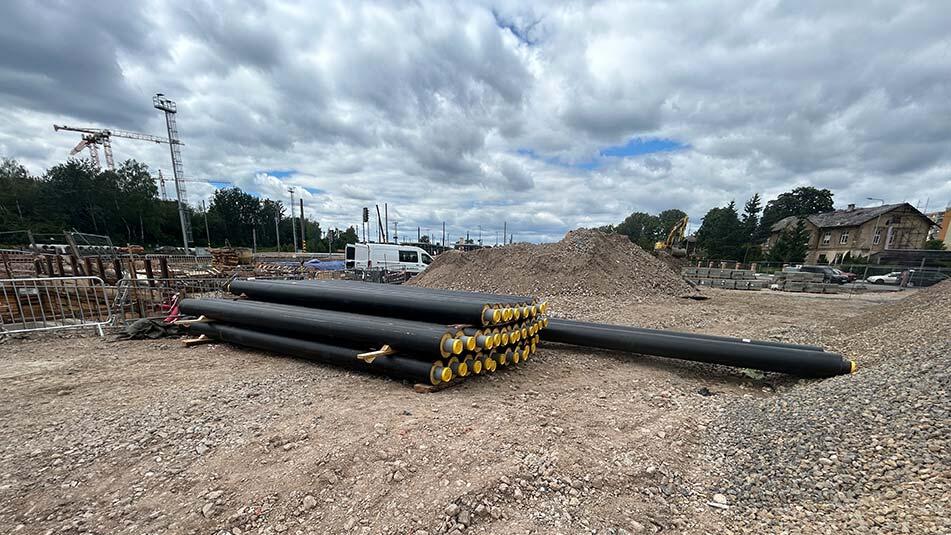 Stack of large black pipes with yellow ends stacked on a gravel construction site, with a dirt mound and crane in the background.