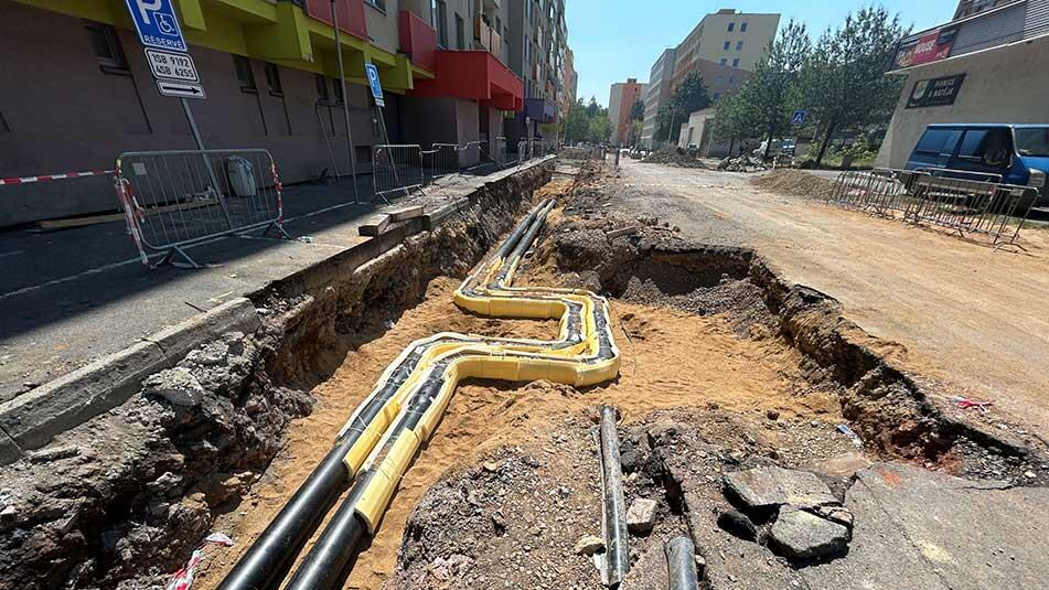 Open trench along a city street with yellow and black utility pipes laid in the dirt and metal barriers nearby, during construction.